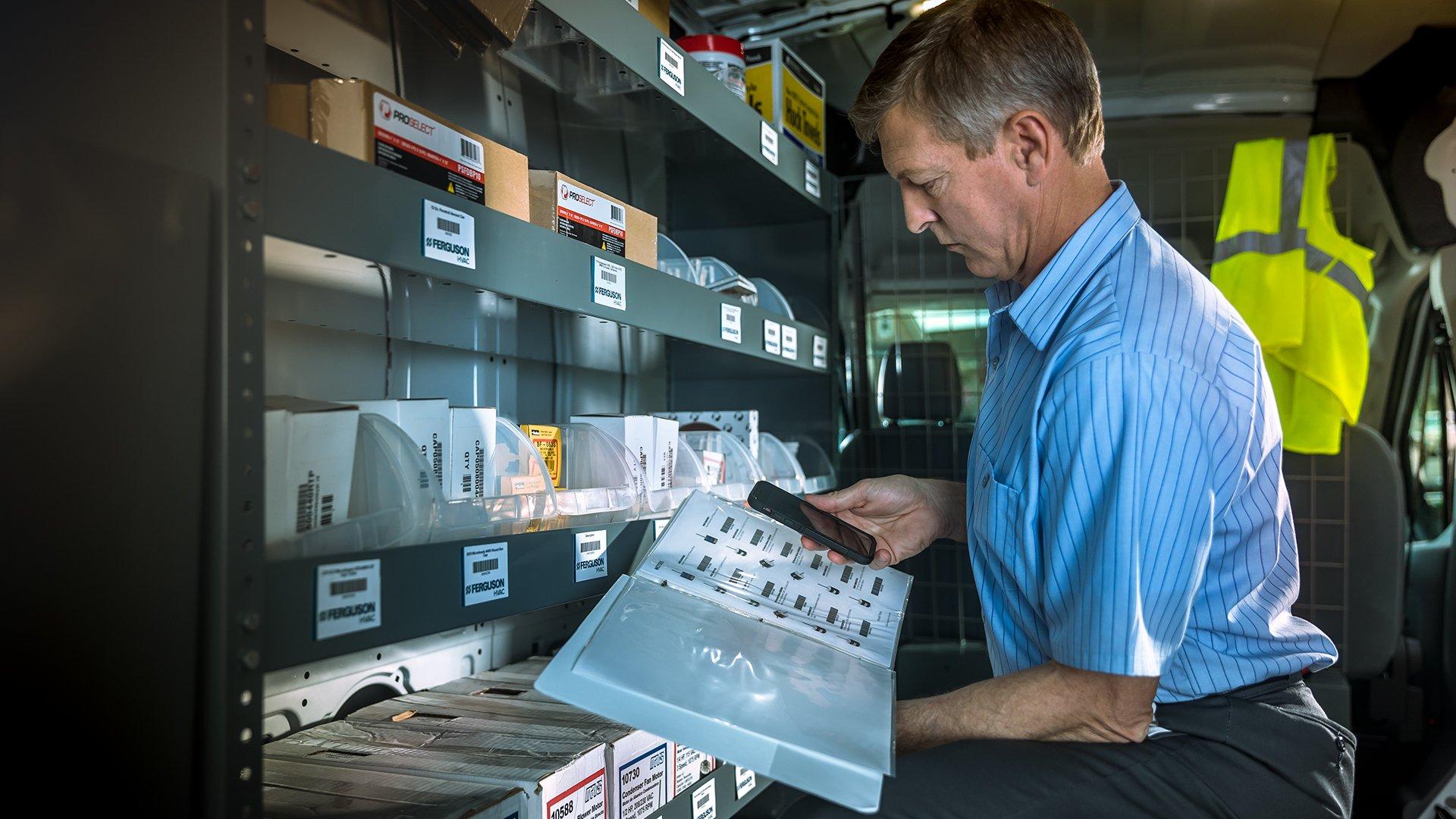 An HVAC technician in his work van uses his phone to scan barcodes of parts in a price book.