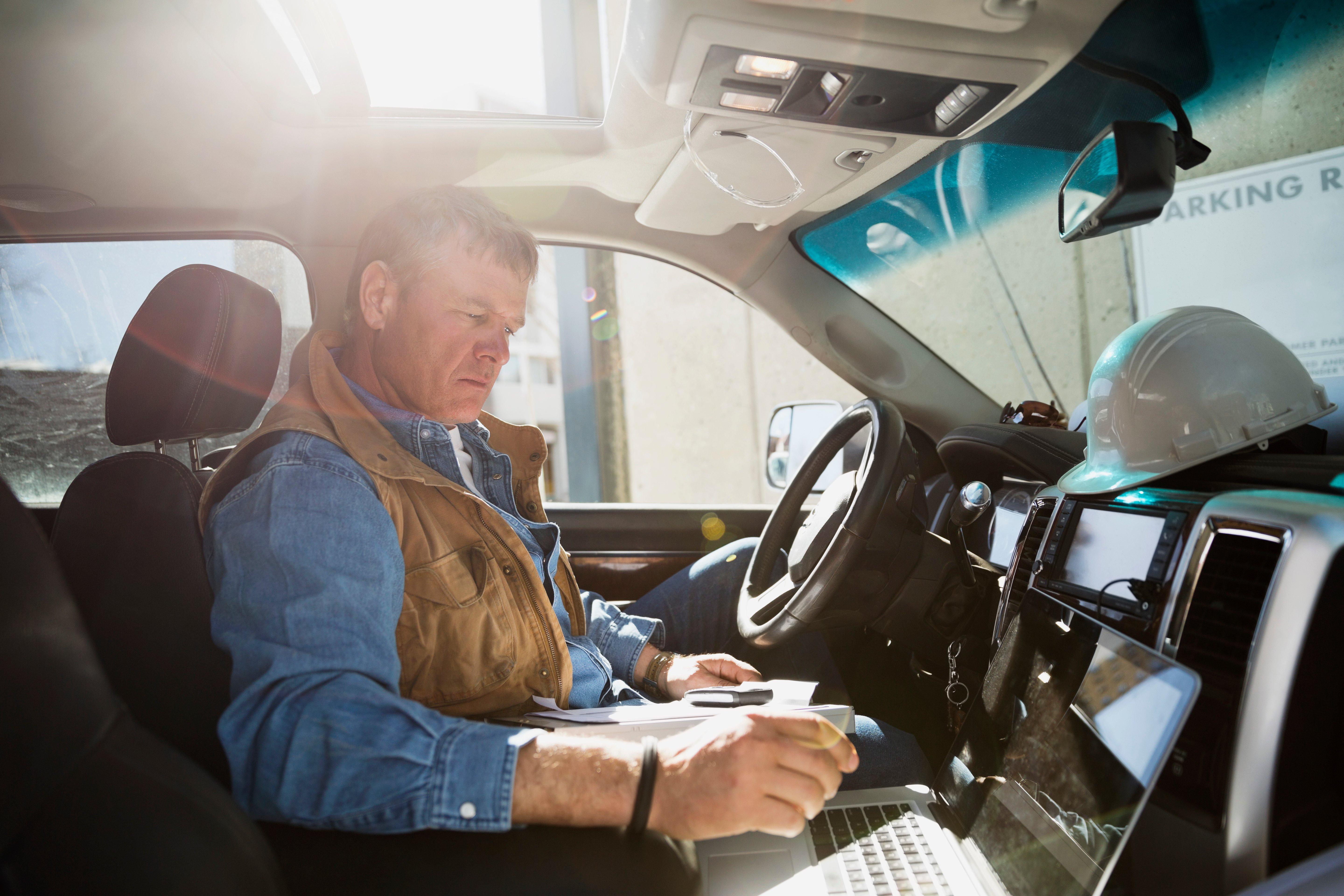 A contractor in the driver's seat of a parked truck reviews his laptop.