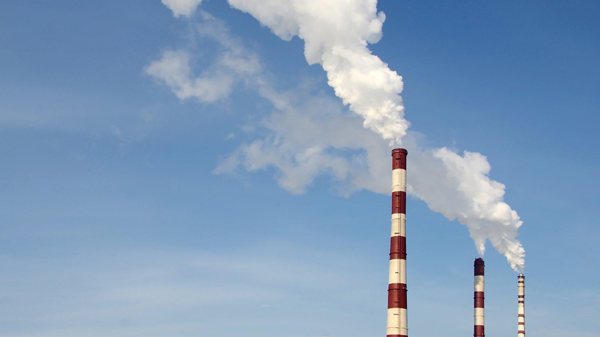 Against a blue sky, white smoke billows out of three red and white industrial chimneys.