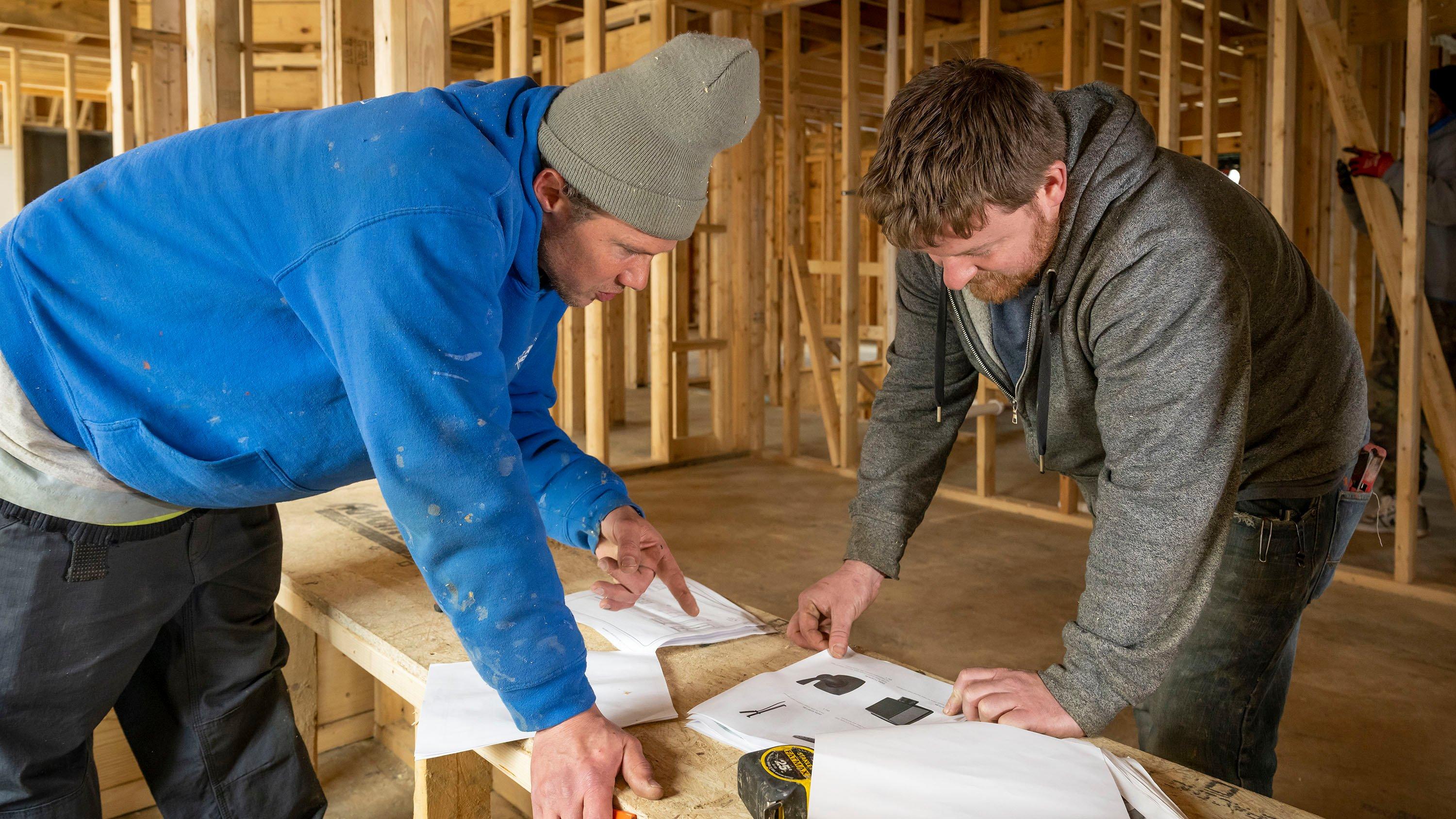 Two men review instruction manual inside framed residential building.