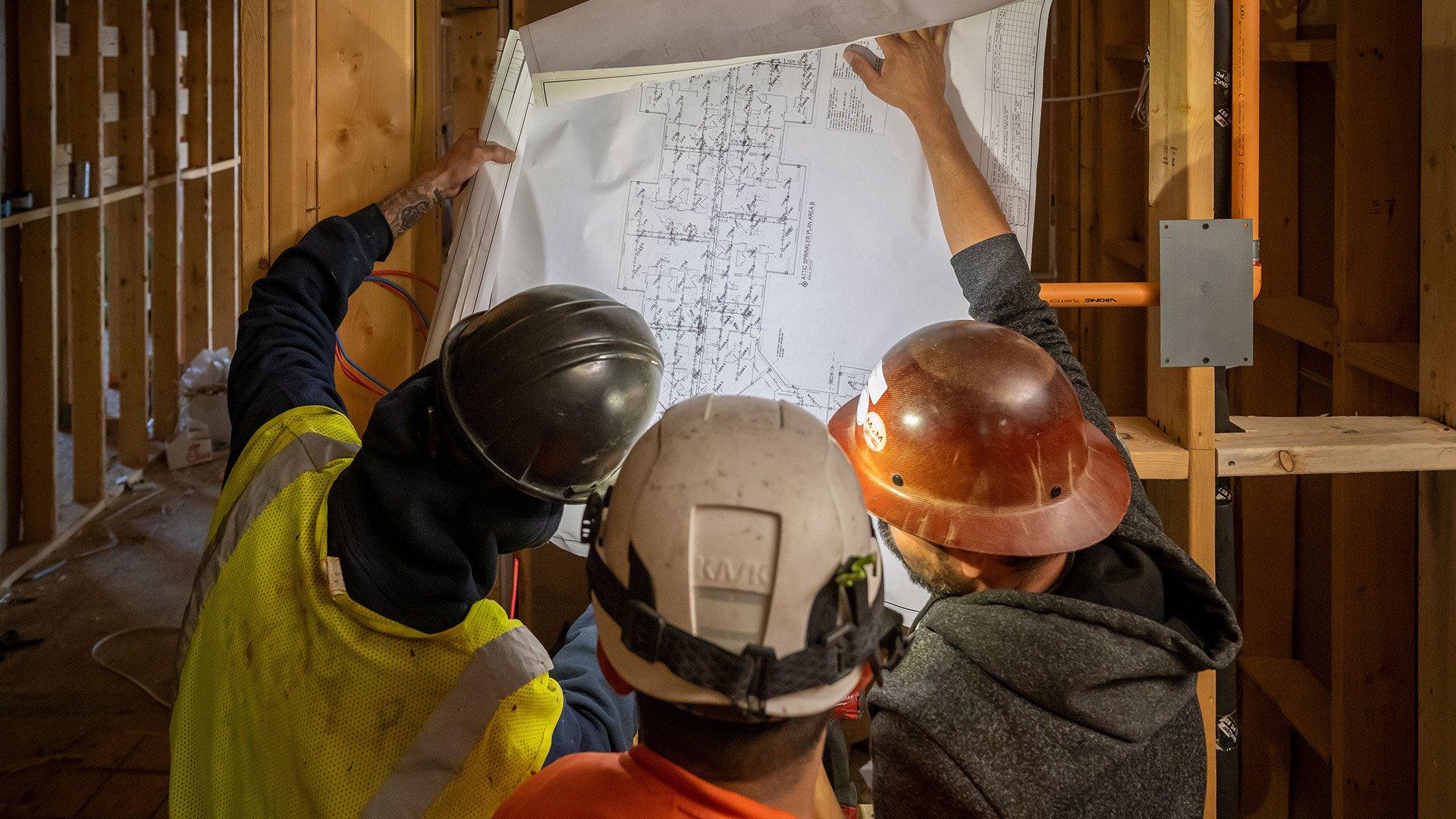 Three individuals in hardhats evaluating a building blueprint on a dark construction site.