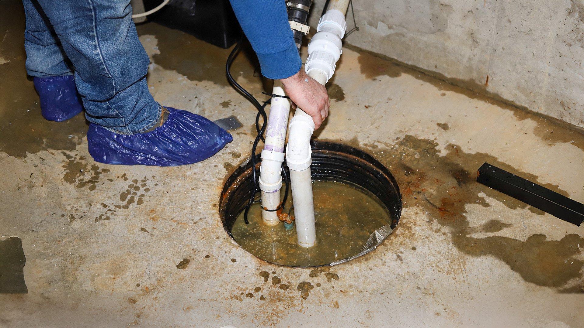 Technician wearing blue disposable shoe covers holds two white PVC pipes that lead into a sump pit in the concrete floor. 