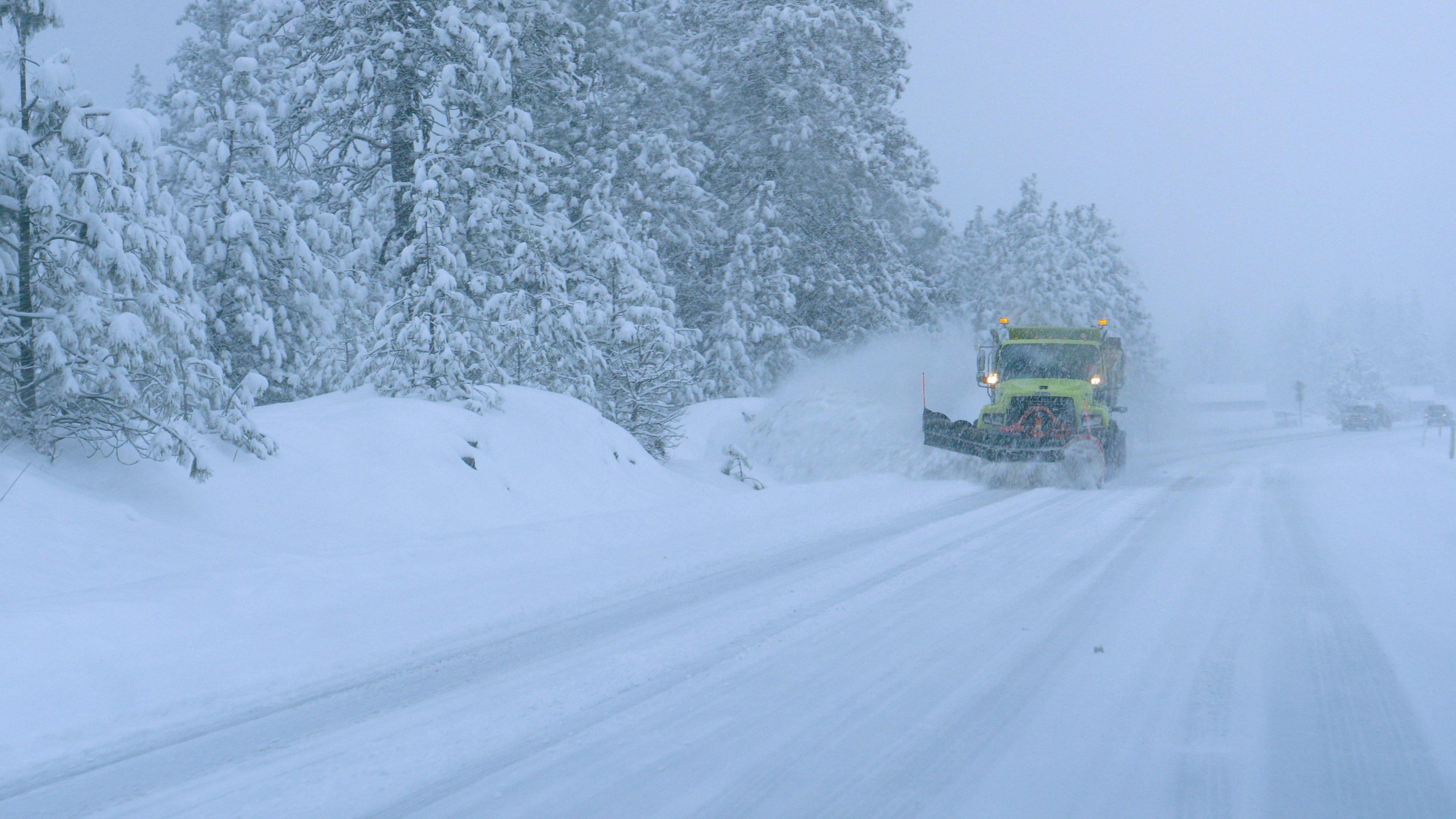 A snow plow cleans up a wintry road next to a forest.