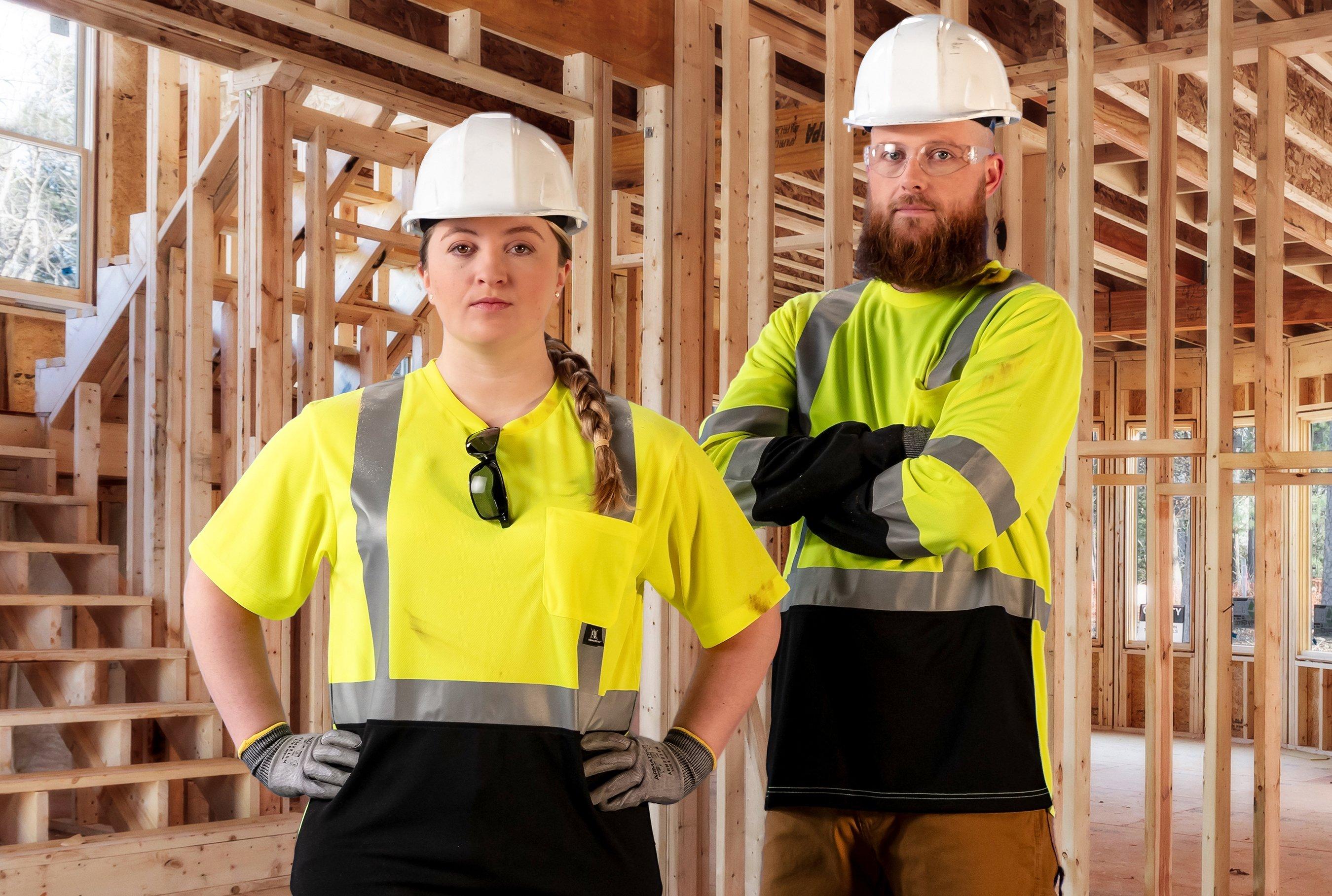Two contractors wearing yellow reflective vests and hard hats stand with their arms crossed in a residential new build.