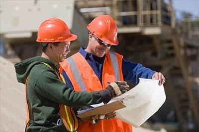 Two workers wearing orange hard hats on a construction jobsite review a blueprint.