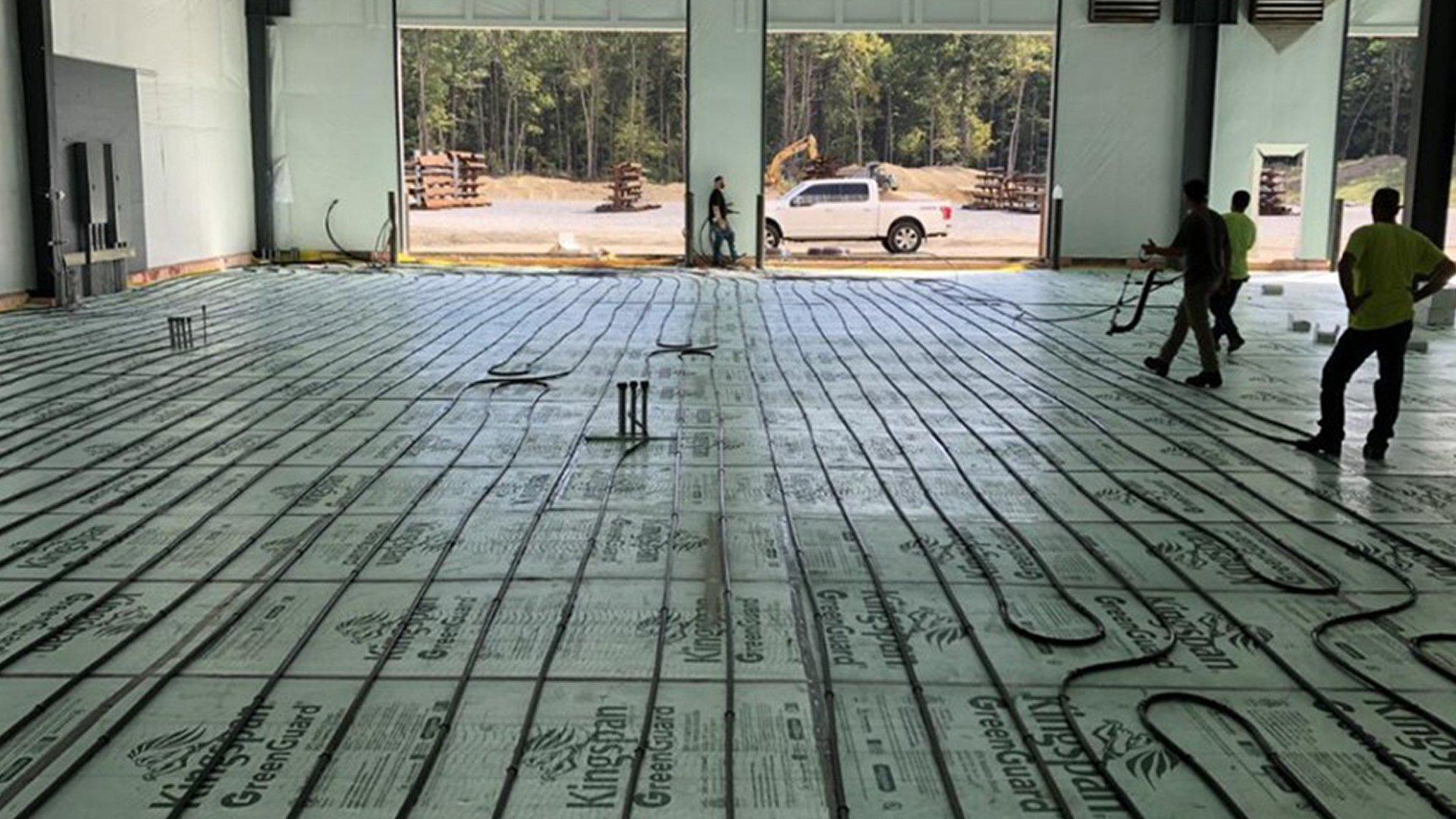 Workers lay out black rubber tubing for radiant floor heating in a warehouse.
