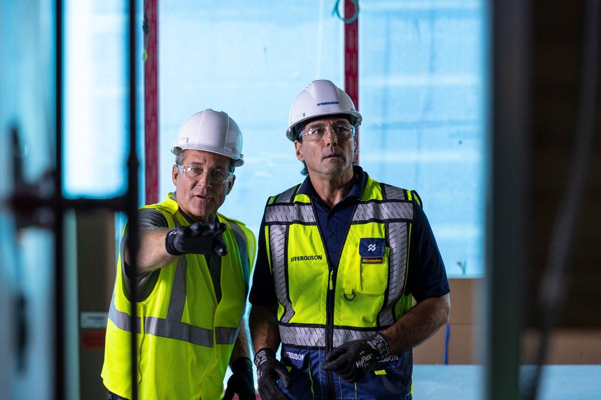 Two Ferguson associates wearing hard hats and reflective vests talk in a distribution center.