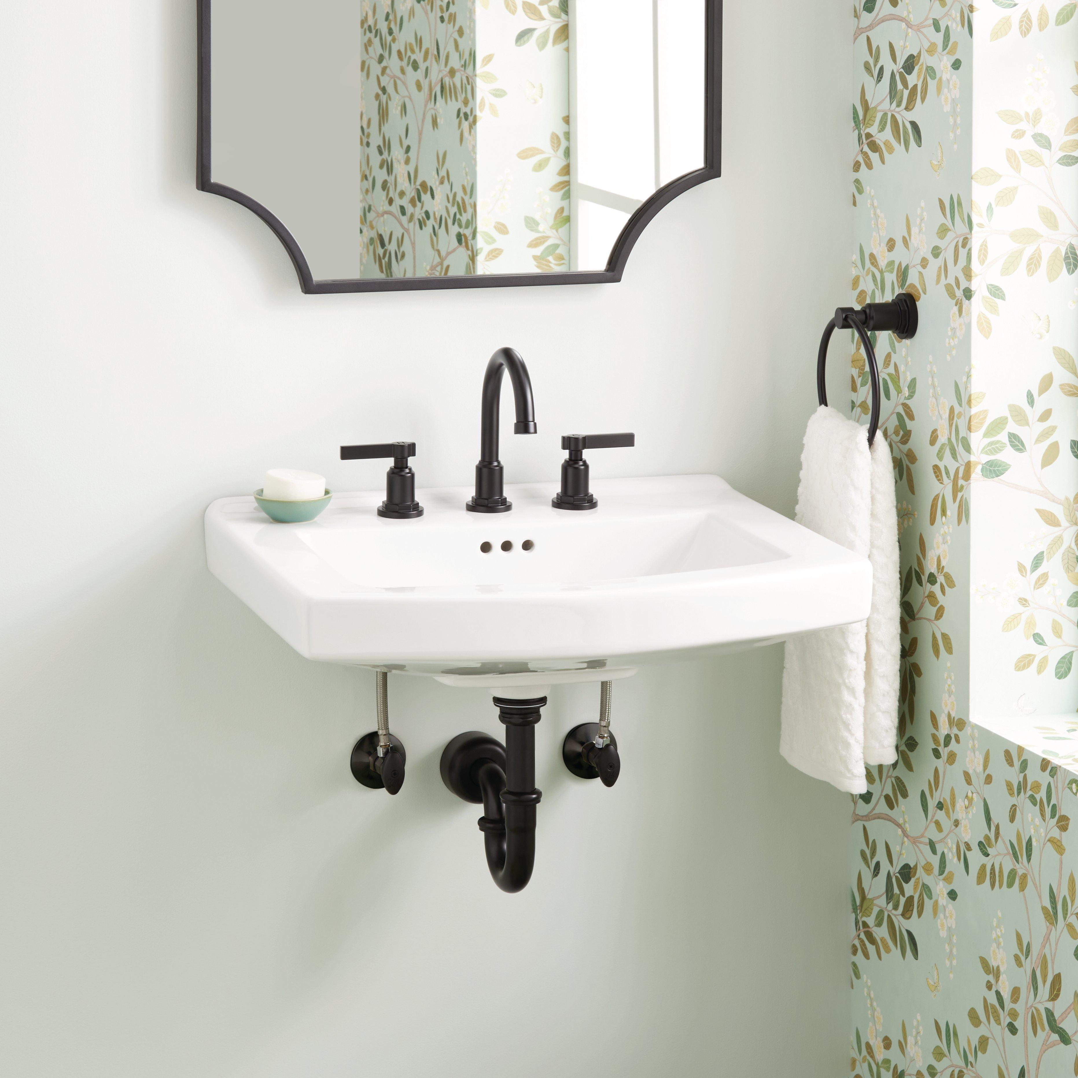 A Pennfield porcelain wall-mounted sink, white with black trim, is the focal point in this guest bathroom with pale green walls, a flowered accent wall and a mirror with a black frame.