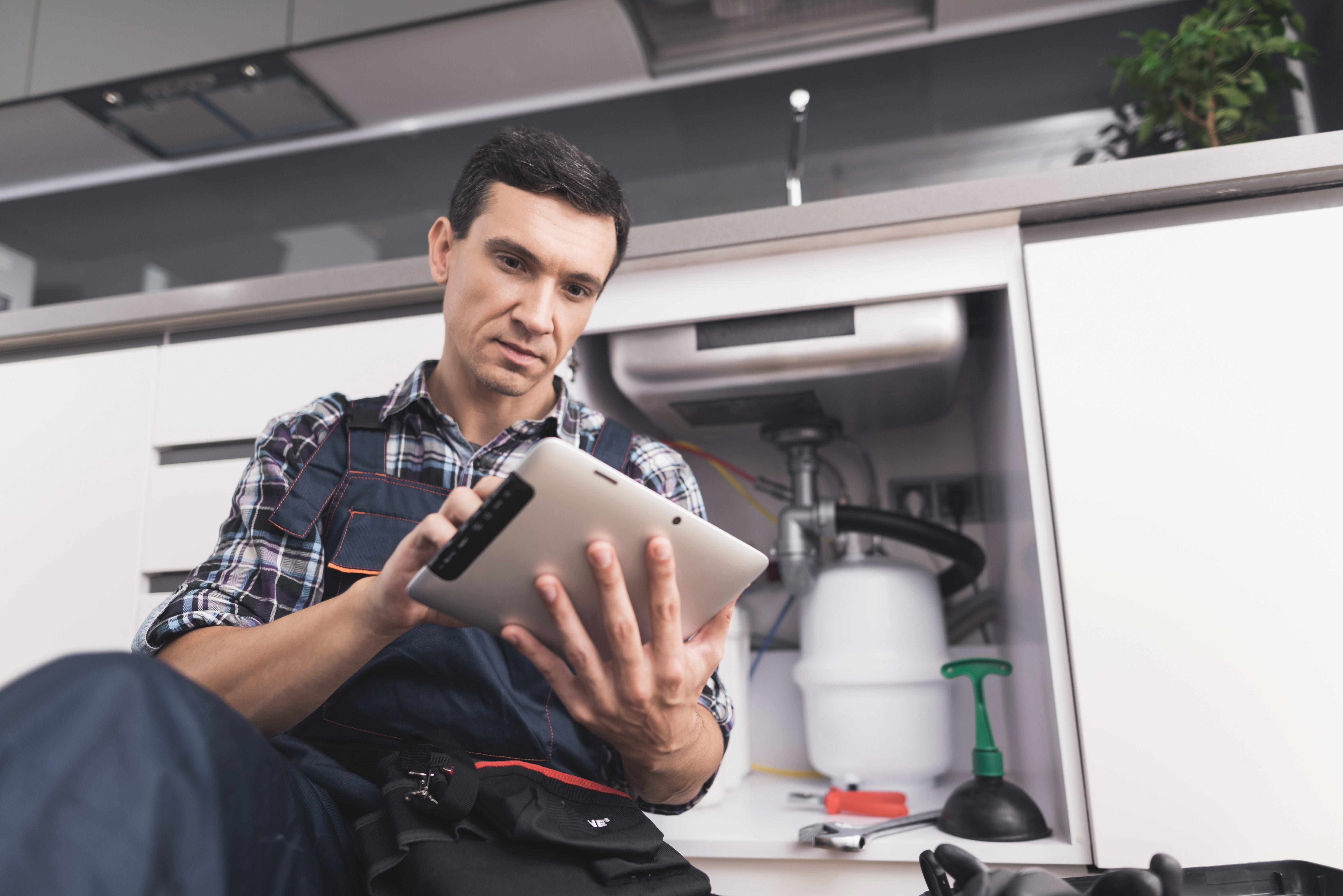 A plumber sits in front of a kitchen sink while he reviews his tablet.