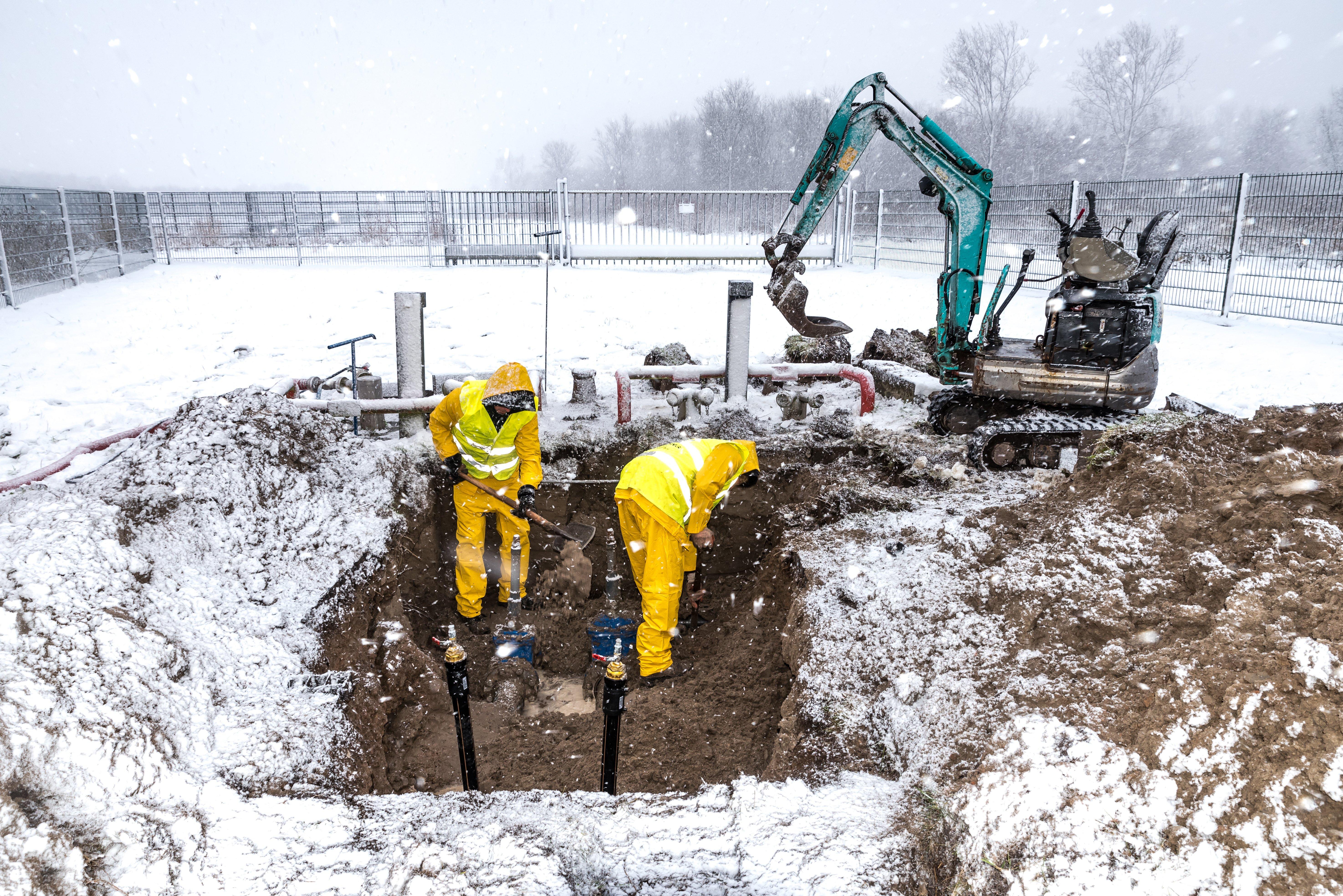 Two workers wearing reflective gear are in a trench outside in the snow repairing a damaged water pipe.