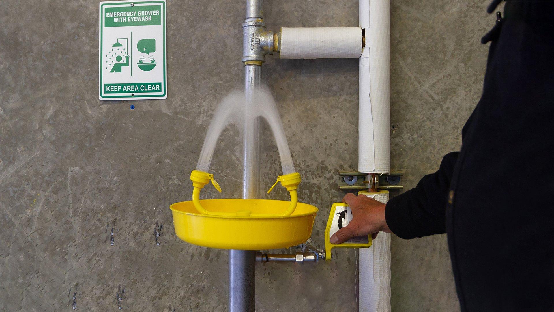 An emergency shower with eyewash water flowing next to a sign inside a facility.