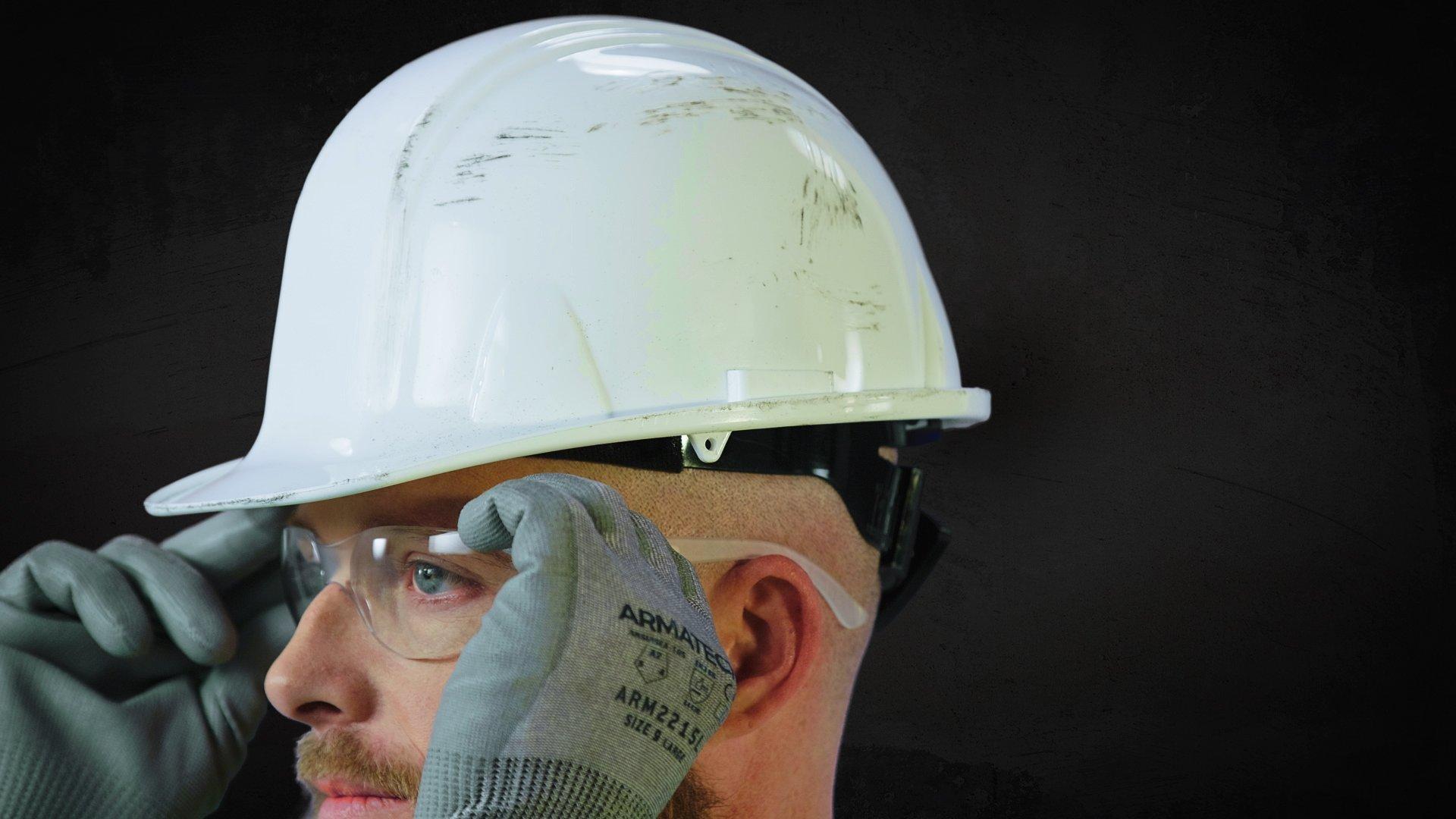 A man wearing a white hard hat and Armateck hand protection puts on safety glasses.