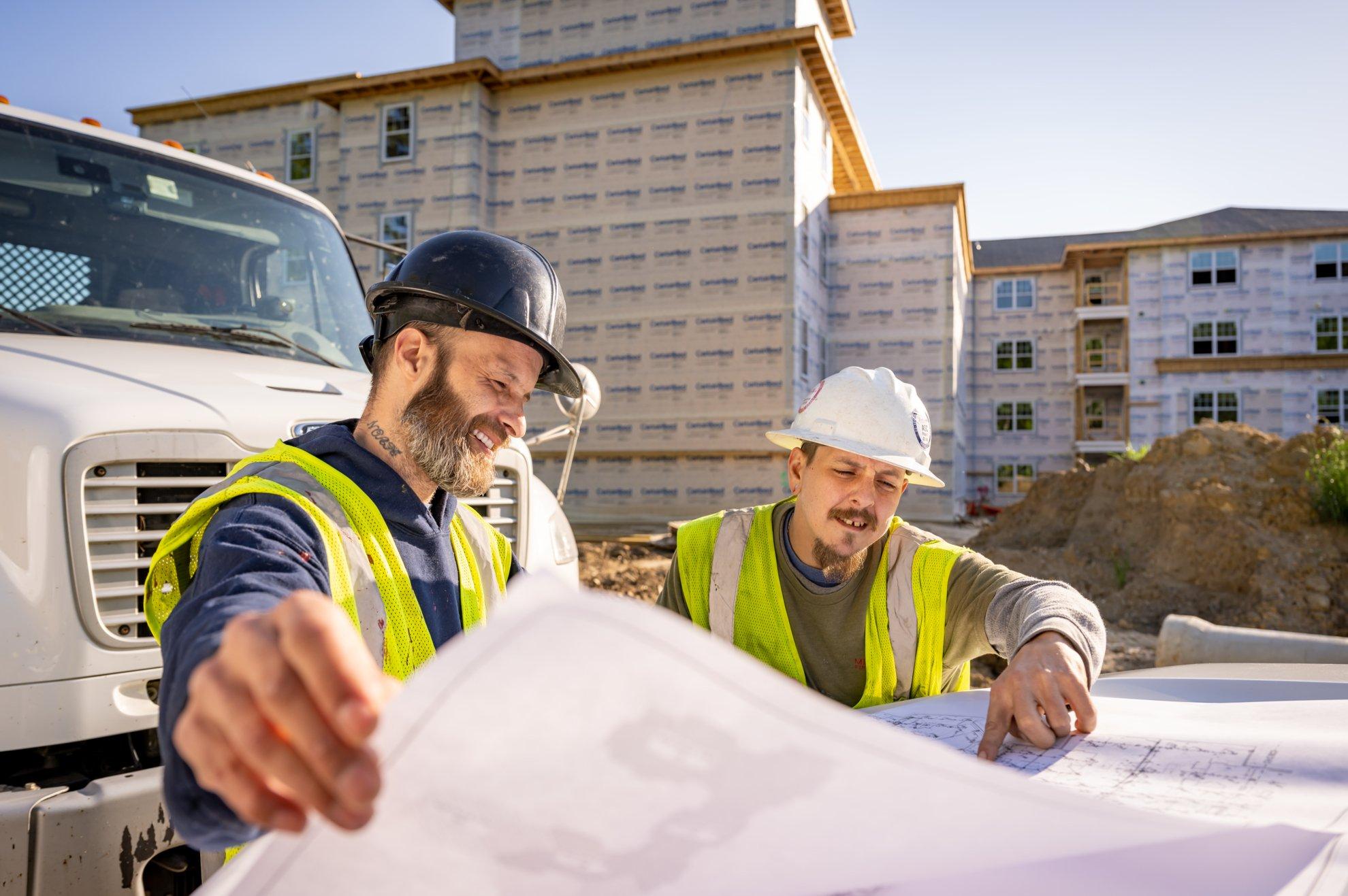 Two workers in hardhats and safety vests stand in front of truck and look at blueprints.