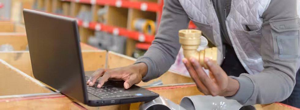 In a warehouse, a person holds a pipe fitting and types on their laptop.