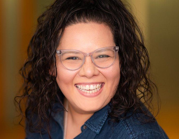 With an orange- and yellow-hued background, a professional headshot depicts a brilliantly smiling woman wearing clear frame glasses. Allie Perez’s wavy black hair is pulled back and falls over the shoulders of her business-style denim button-down shirt.