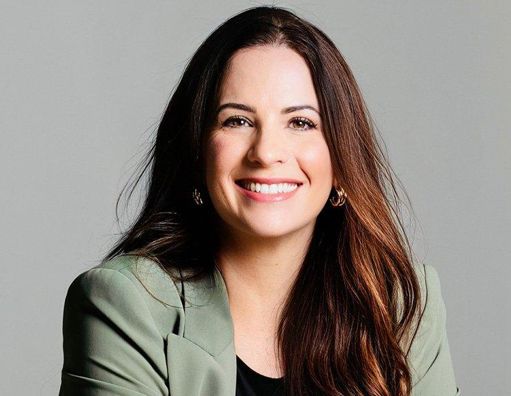 With a light gray background, a professional headshot depicts a woman with long brown hair with a straightforward smile. Julene Arregui appears to lean slightly forward in a welcoming manner and wears a soft green blazer over a black top.