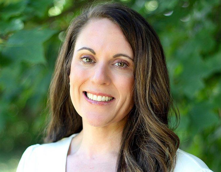 Against a background of green, leafy trees, Katie Byrd smiles in this professional headshot. Her dark brown hair is parted on her right and falls over her left shoulder, and she is wearing a white business-style shirt.