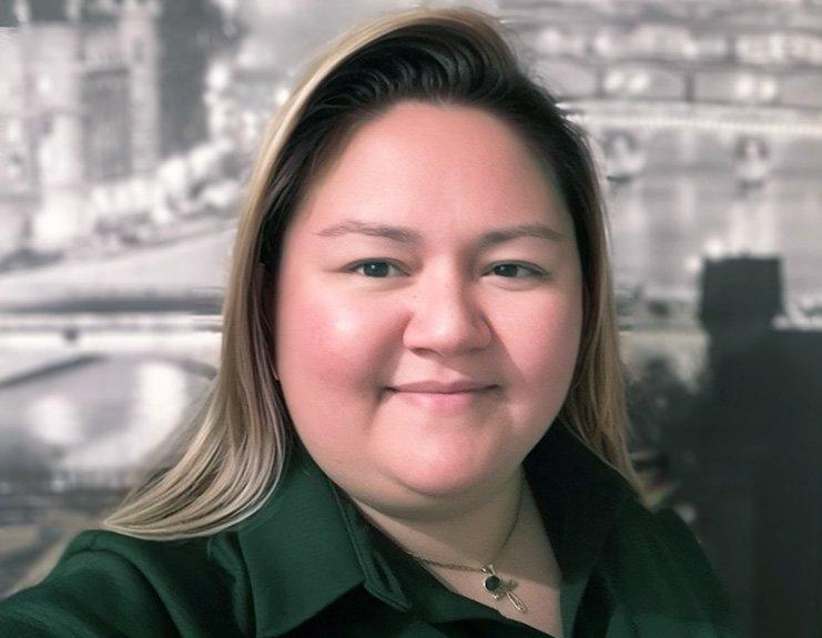 In a professionally taken selfie in front of a cityscape, Kayley Villegas wears a forest green button-down shirt and smiles. Her hair has blonde layers and is parted on her left side.