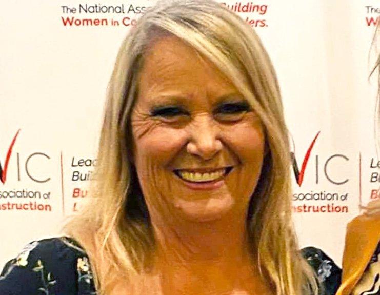 In front of a NAWIC step and repeat banner, Linda Cudmore smiles broadly. Her blonde hair is parted on the right side and falls over her shoulders. She wears a black flowered top.