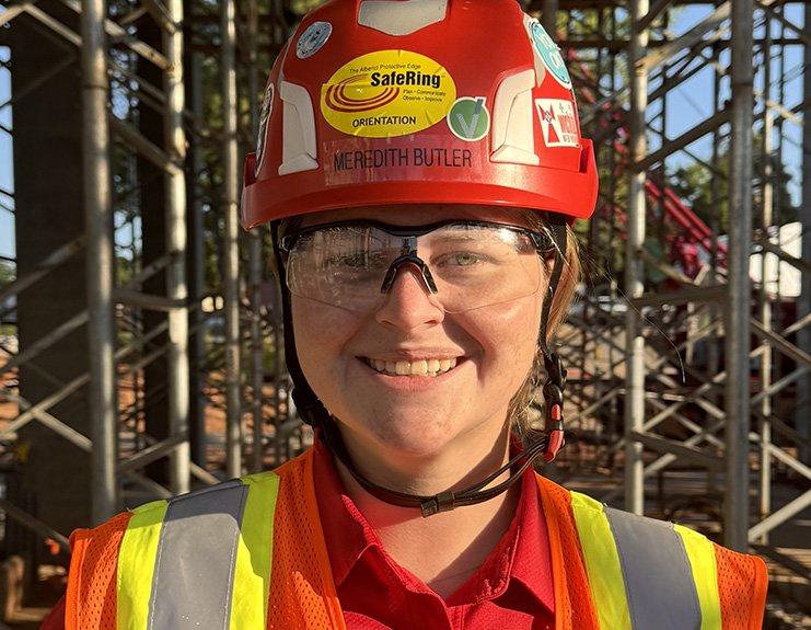 At a worksite with scaffolding behind her, Meredith Butler smiles. She wears a red hard hat with her name on it, safety goggles, and a yellow reflective vest over a red button-down shirt.