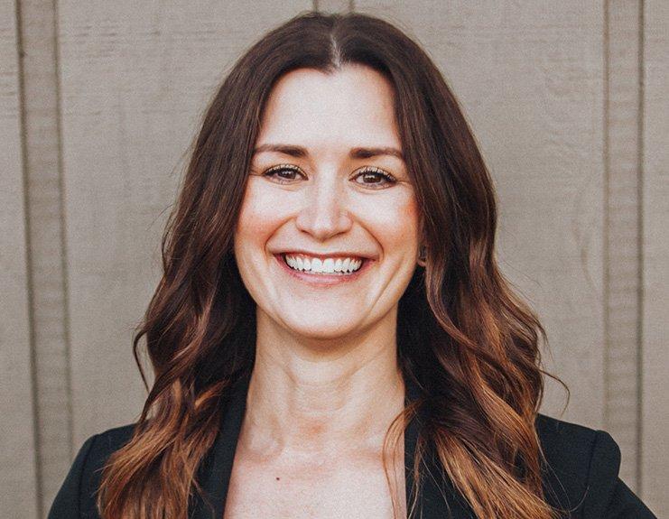 In front of a light-colored wooden background, Rachel Duggan smiles brightly. Her long, wavy brown hair is parted in the middle and she wears a black blazer.