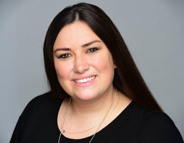 With a light grey background, this professional headshot depicts Renee Lenox learning forward and smiling. Her long black hair is parted off center and she wears a long-sleeve black shirt.