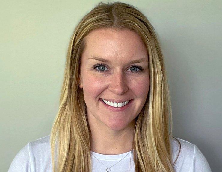 Against a light-colored wall, Sarah Smith smiles. Her long blonde hair is parted just off center and she is wearing a white jersey.