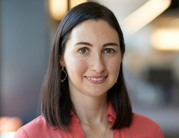 In front of a blurred office setting, Shelly Kalb smiles into the camera. Her dark brown hair is parted on the left and falls to just beyond her shoulders. She is wearing a salmon-colored button-down shirt.