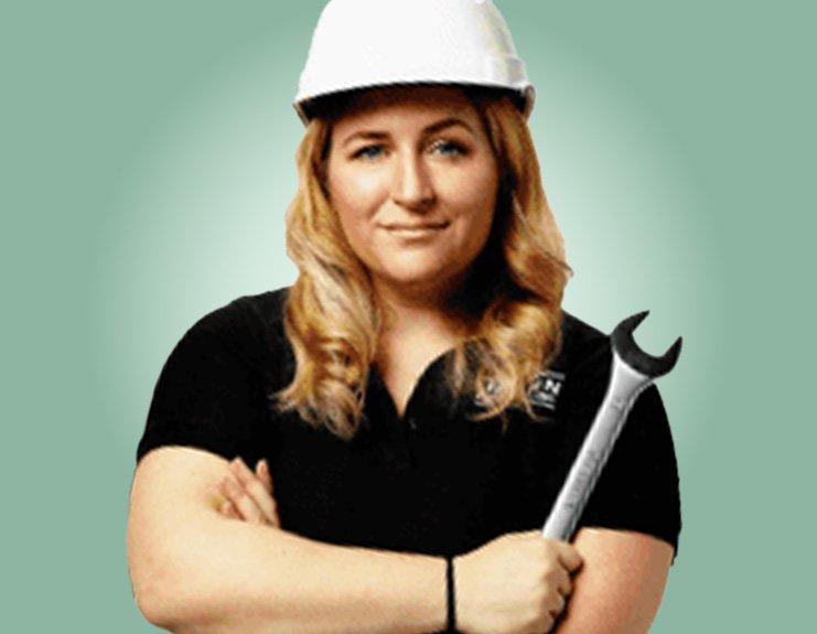 In this professional headshot against a light mint-green background, Skylar Halpin wears a white hard hat, a black business T-shirt with her company’s logo, and carries a wrench in her crossed arms.