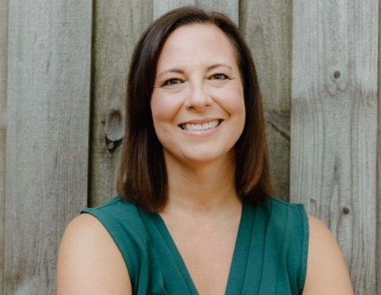 In front of a wooden privacy fence, Stephanie Hadley smiles warmly. Her dark brown hair is parted on the left and falls to her shoulders, and she is wearing a sleeveless dark-green business top.