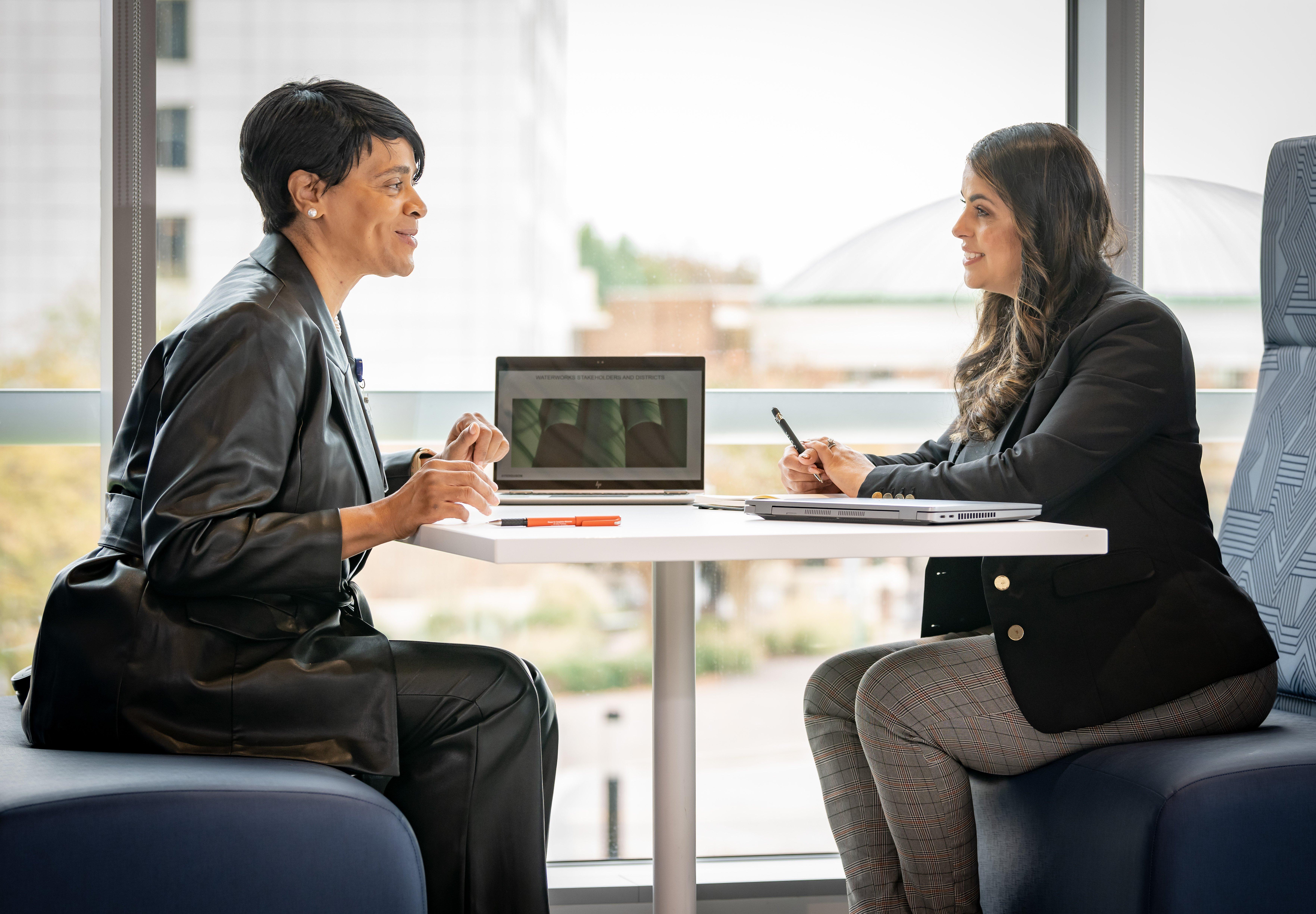 Two women in professional clothing talk with each other at a table with two laptops and notepads.