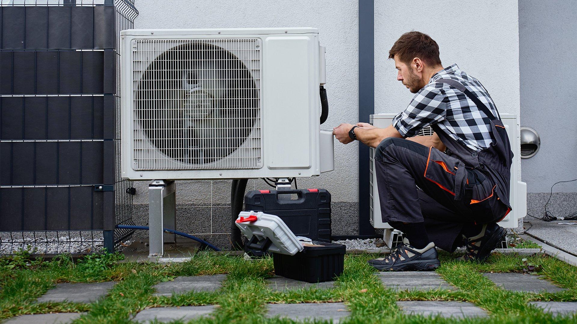 HVAC technician kneels beside outdoor heat pump unit and inspects. 