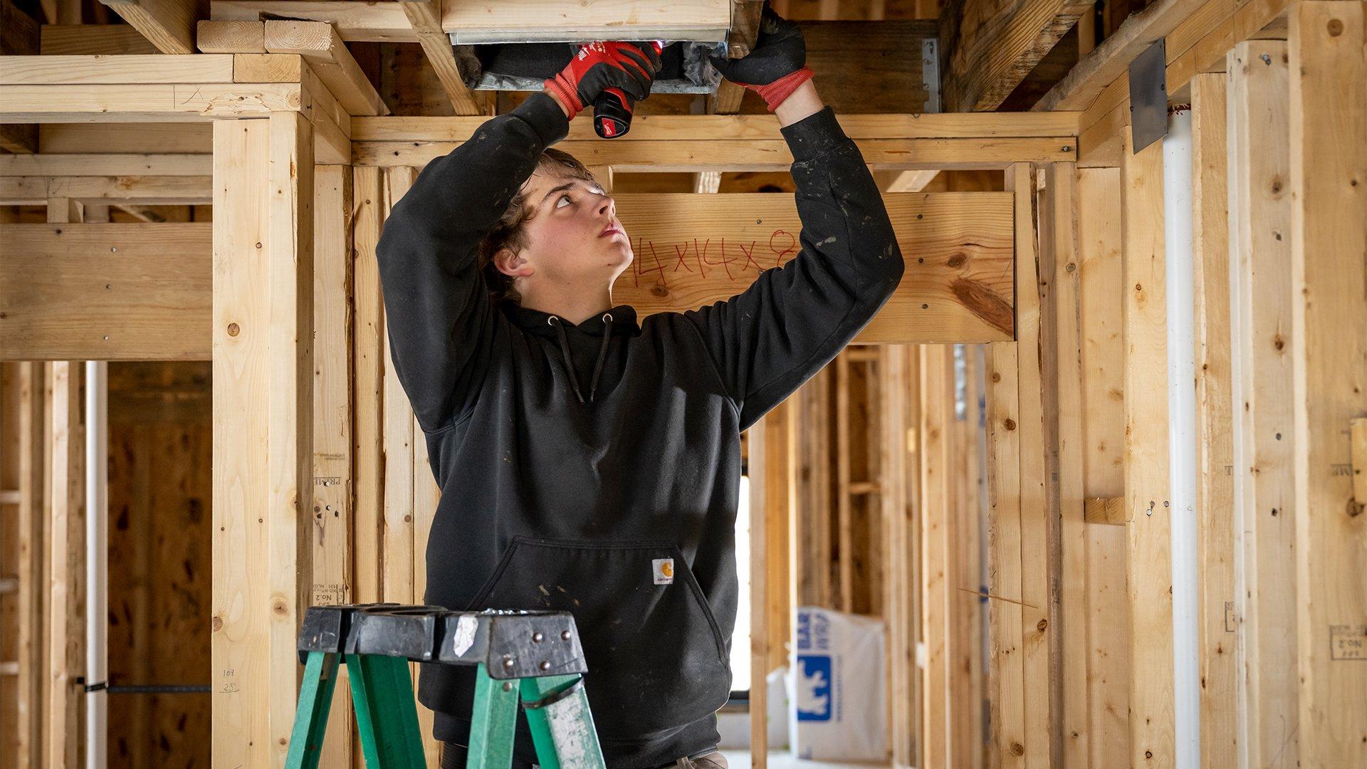 HVAC technician stands on ladder in new residential build and installs duct work. 