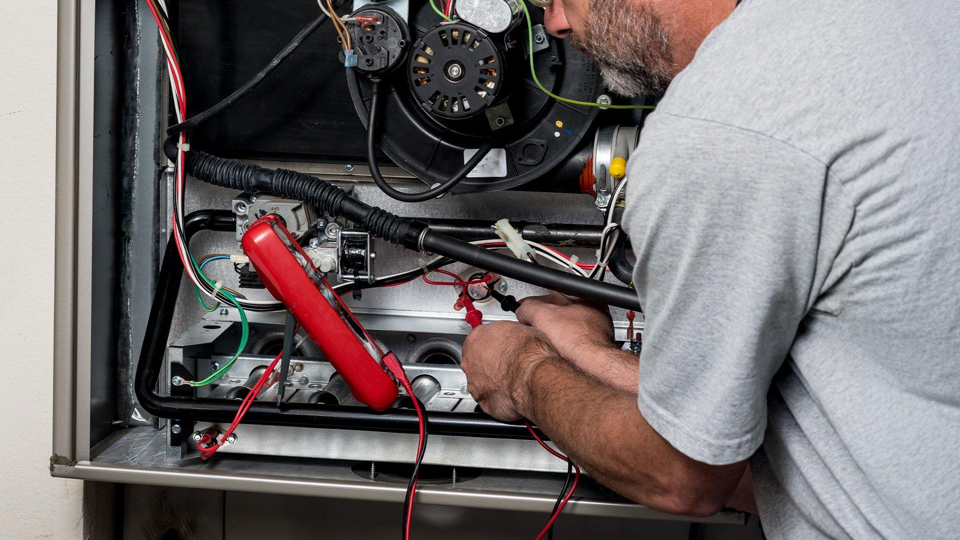 HVAC technician in grey t-shirt uses multimeter on furnace wiring. 
