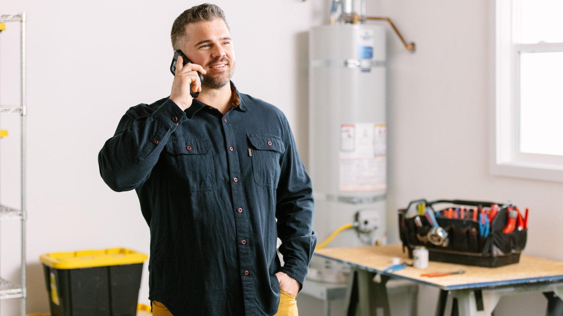 Plumbing contractor talks on phone inside residential garage with water heater in background. 