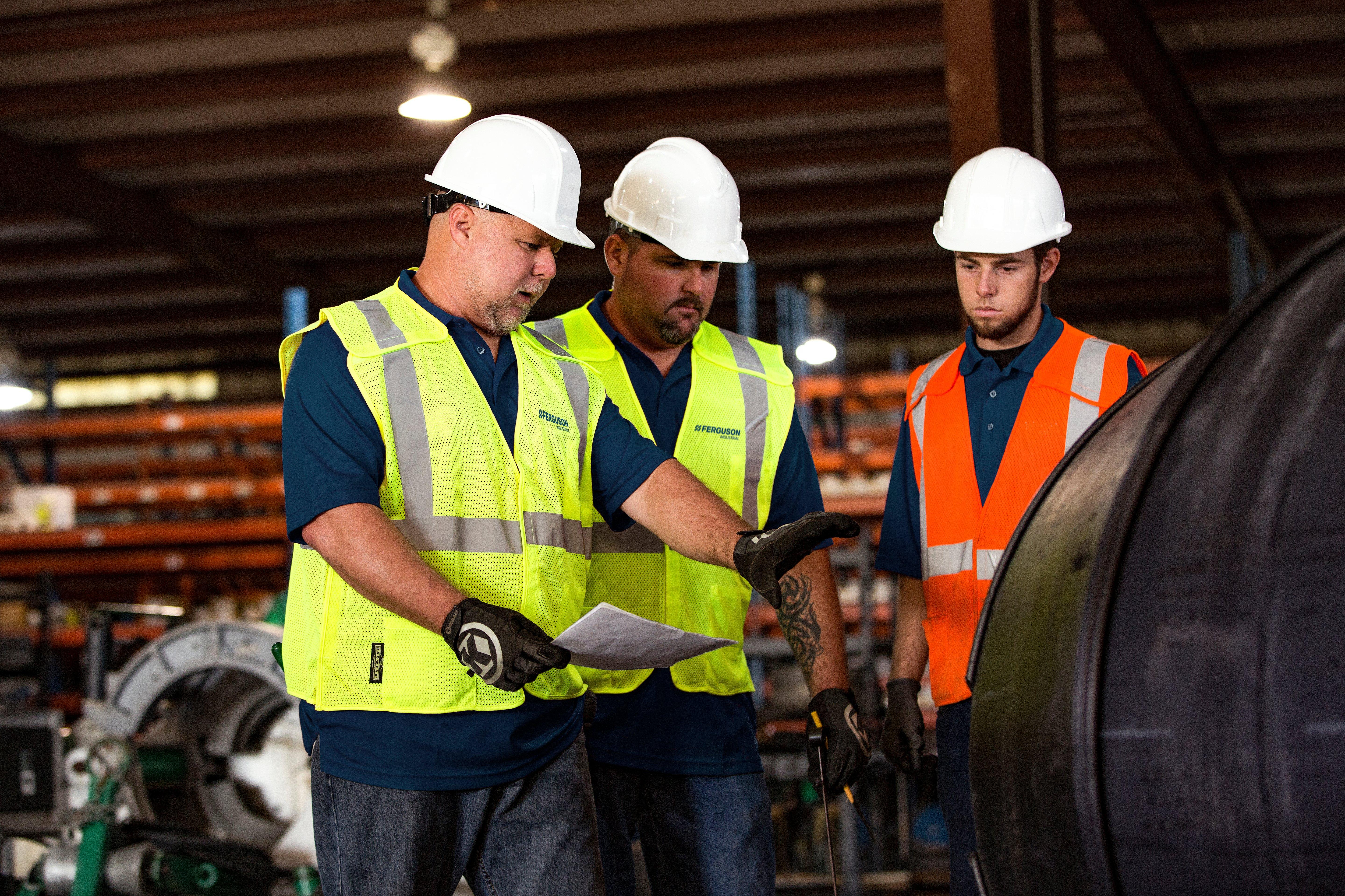 Three Ferguson associates wearing hard hats and reflective vests talk together in front of a large pipe.