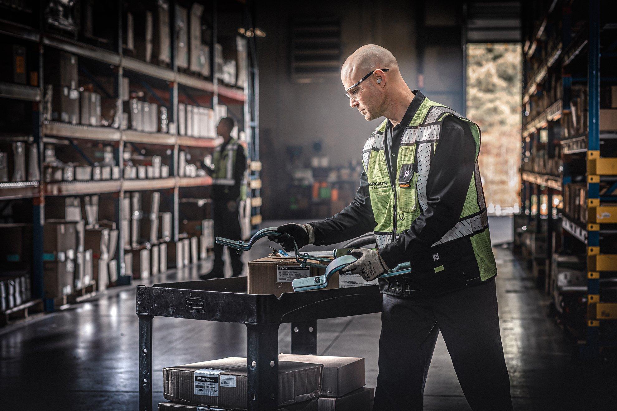 Ferguson associate wearing a reflective vest examines an order of pipe brackets on a wheeled utility cart in a warehouse.