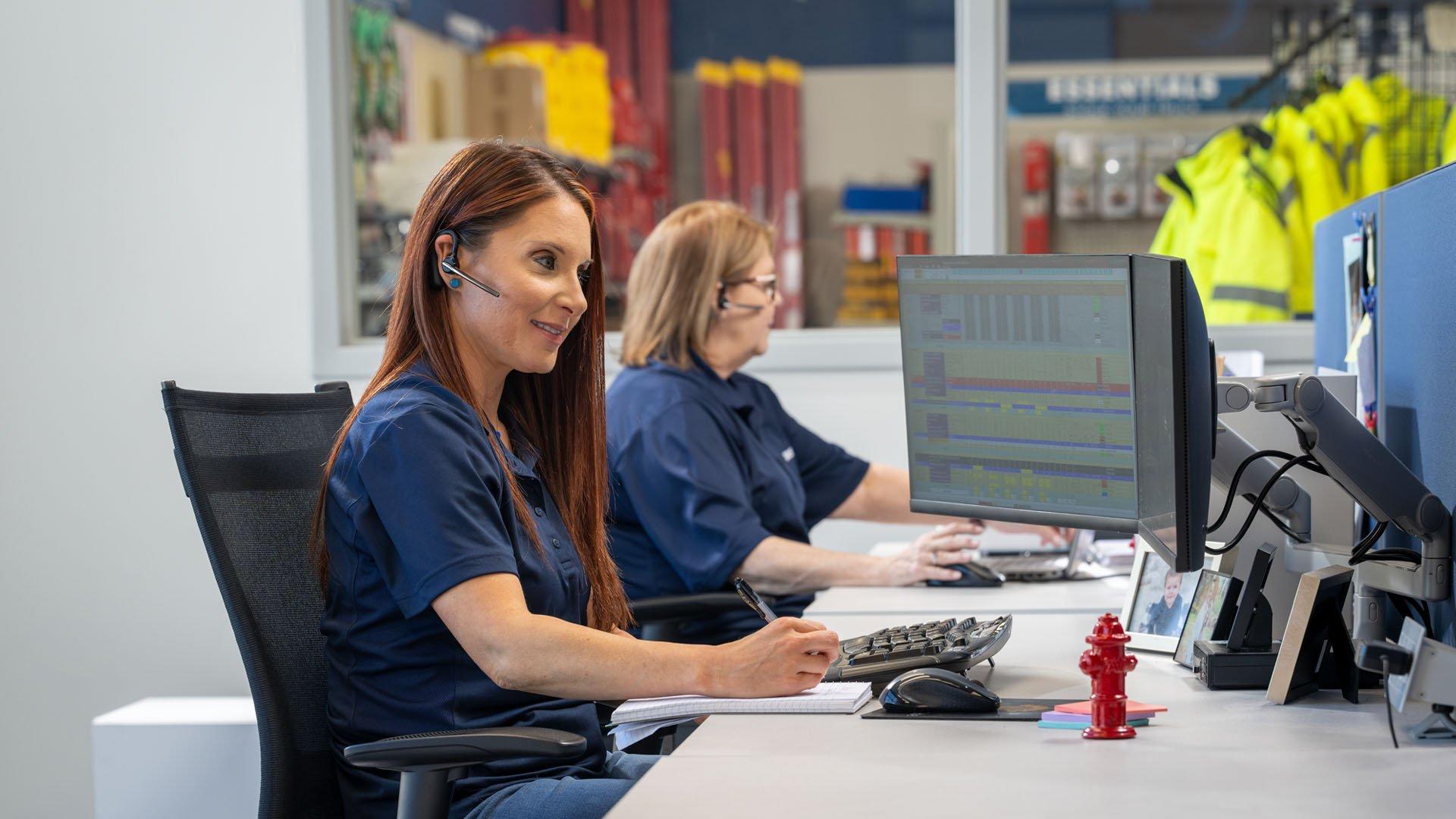 Representatives wearing navy blue shirts, using wireless headsets, sitting in front of computer screens in a call center.