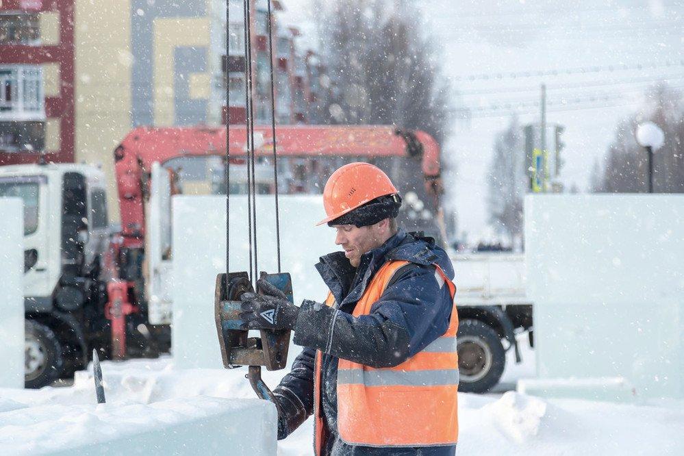 On a snowy city street, a worker wearing an orange reflective hard hat and vest attaches a hook to a beam.