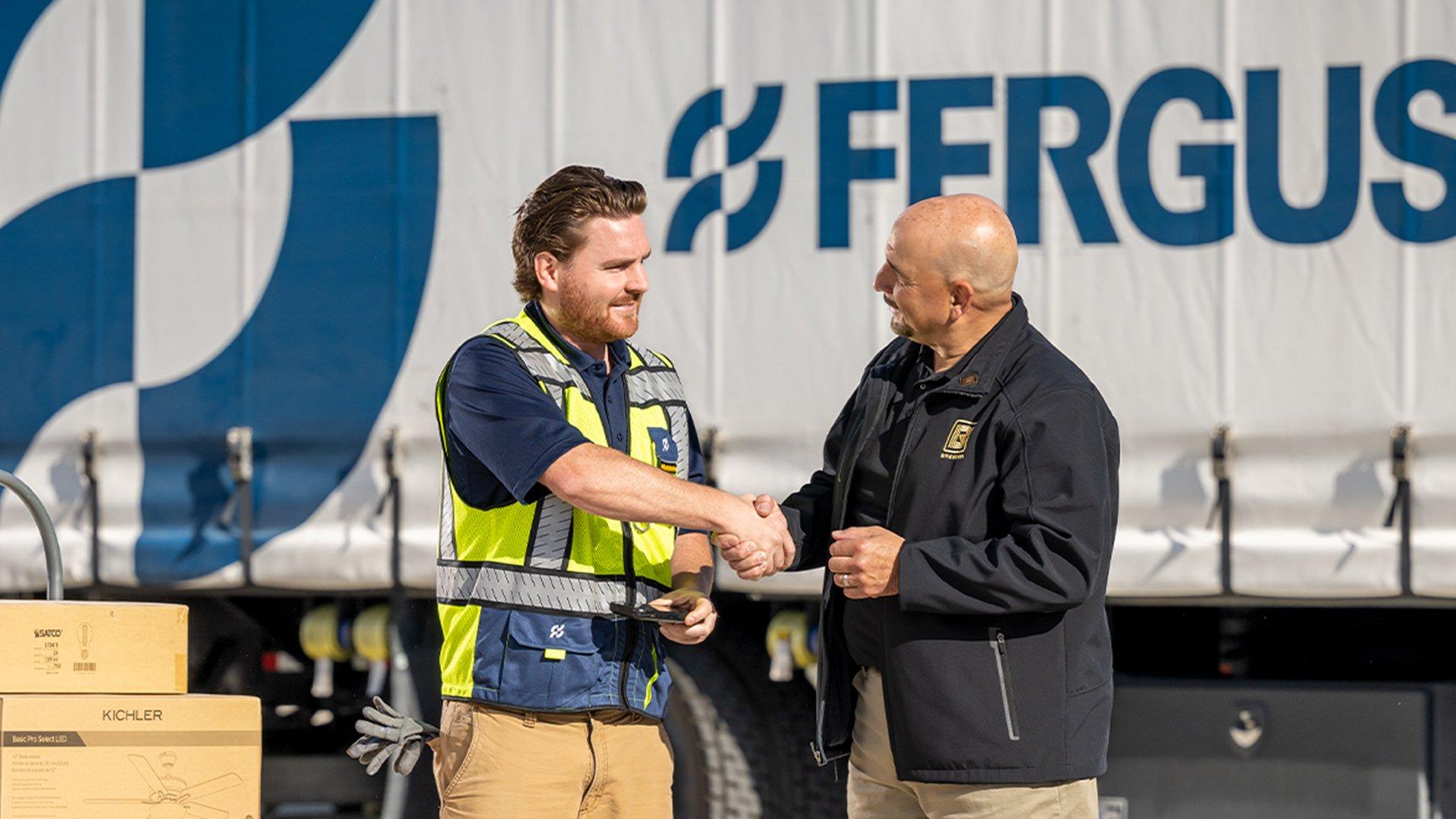 A Ferguson associate shakes hands with a facilities manager next to a stack of boxes on a dolly to their right and a delivery truck parked in the background.