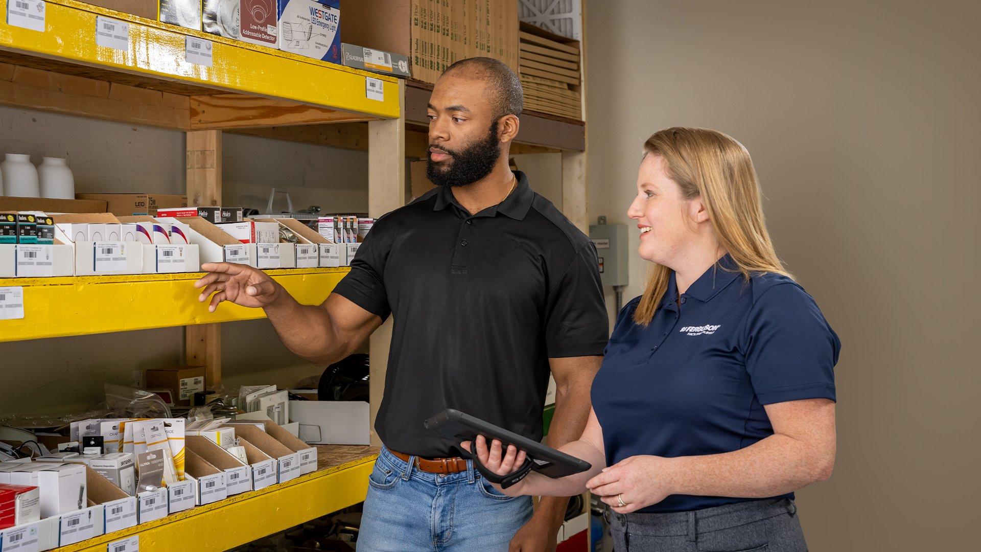 Two individuals evaluate the inventory available on the yellow shelves in their stock room, with one individual holding a tablet an the other pointing at specific items.