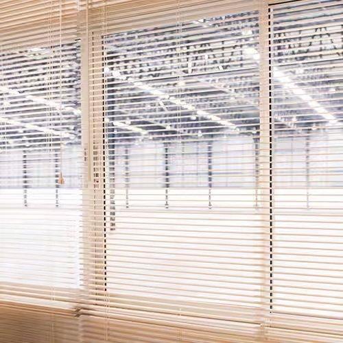 View into a warehouse from an office through open 1-inch white PVC mini blinds.