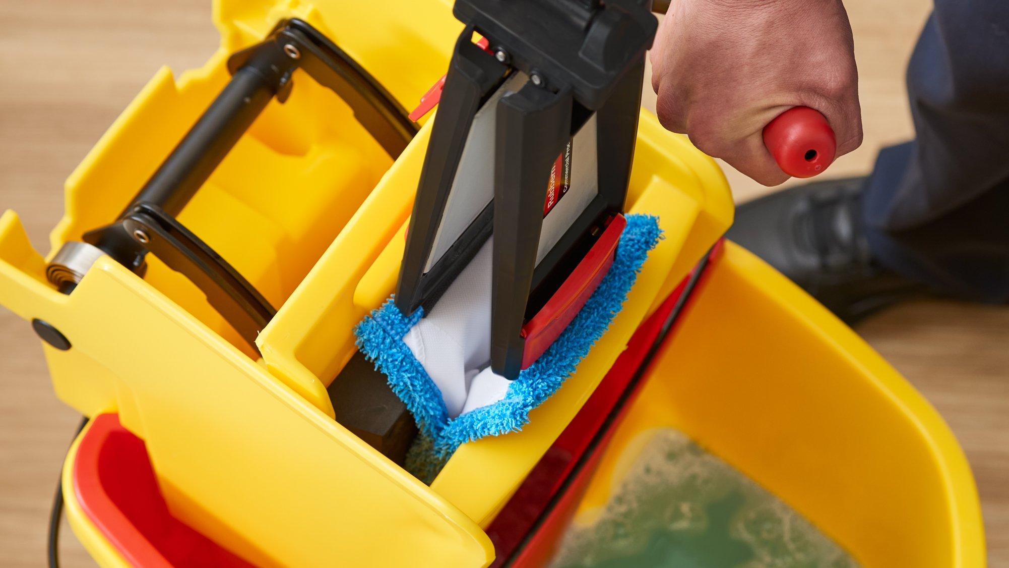 A facilities worker wrings out a flat mop in a yellow bucket.