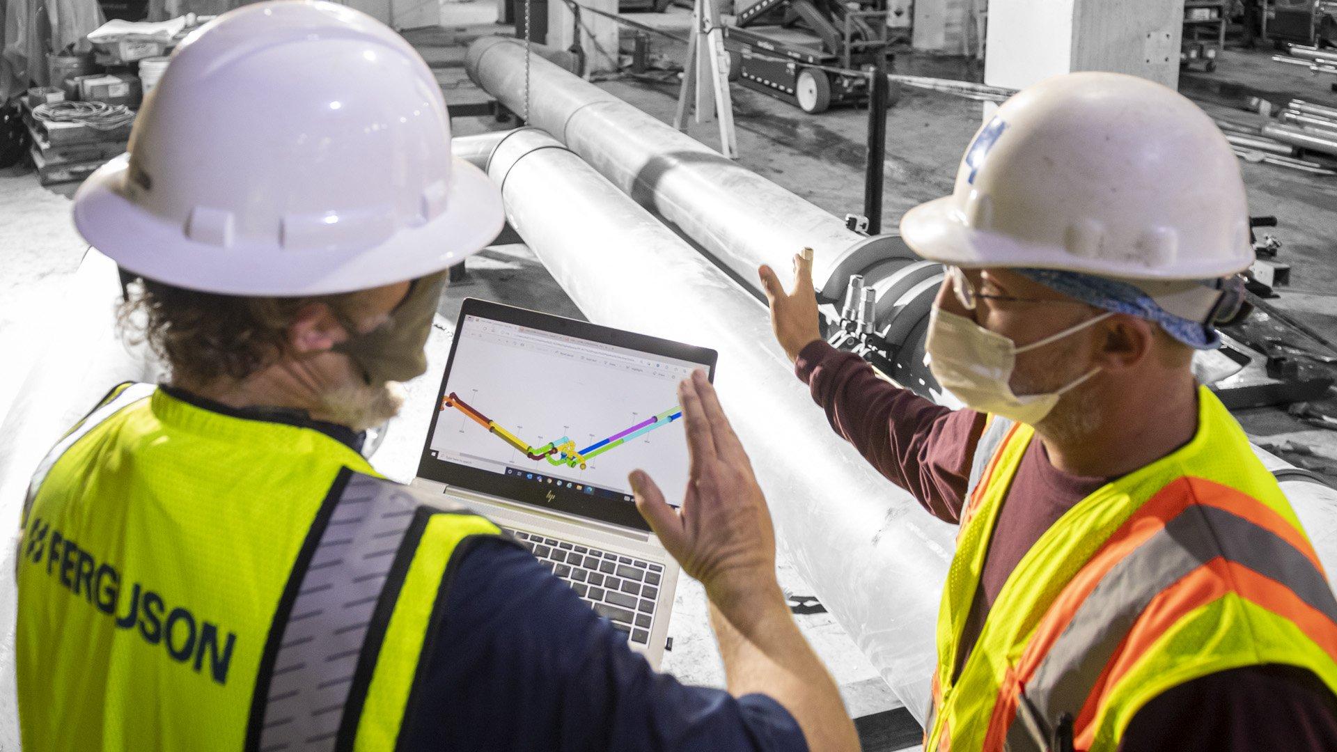 2 associates wearing masks and hard hats converse over laptop with digital services. 