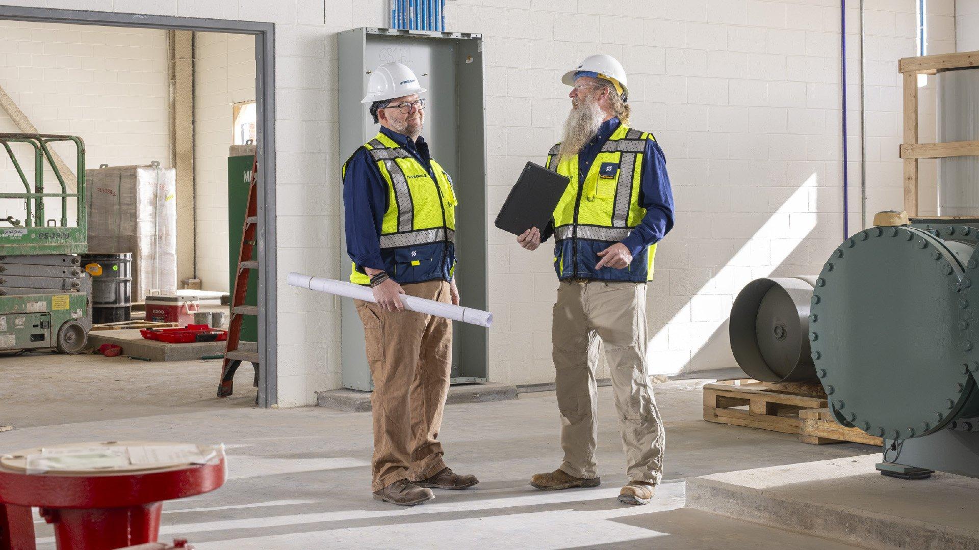 Two associates with hard hats and safety vest talk inside a construction jobsite. 
