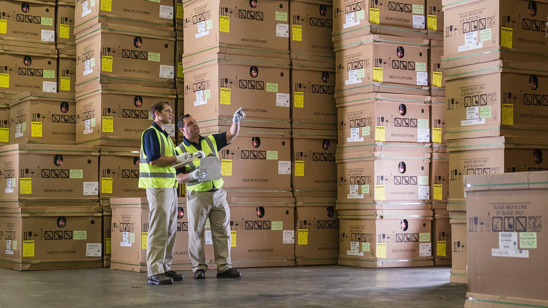 Two warehouse employees with safety vests stand beside product boxes and point to off camera object. 