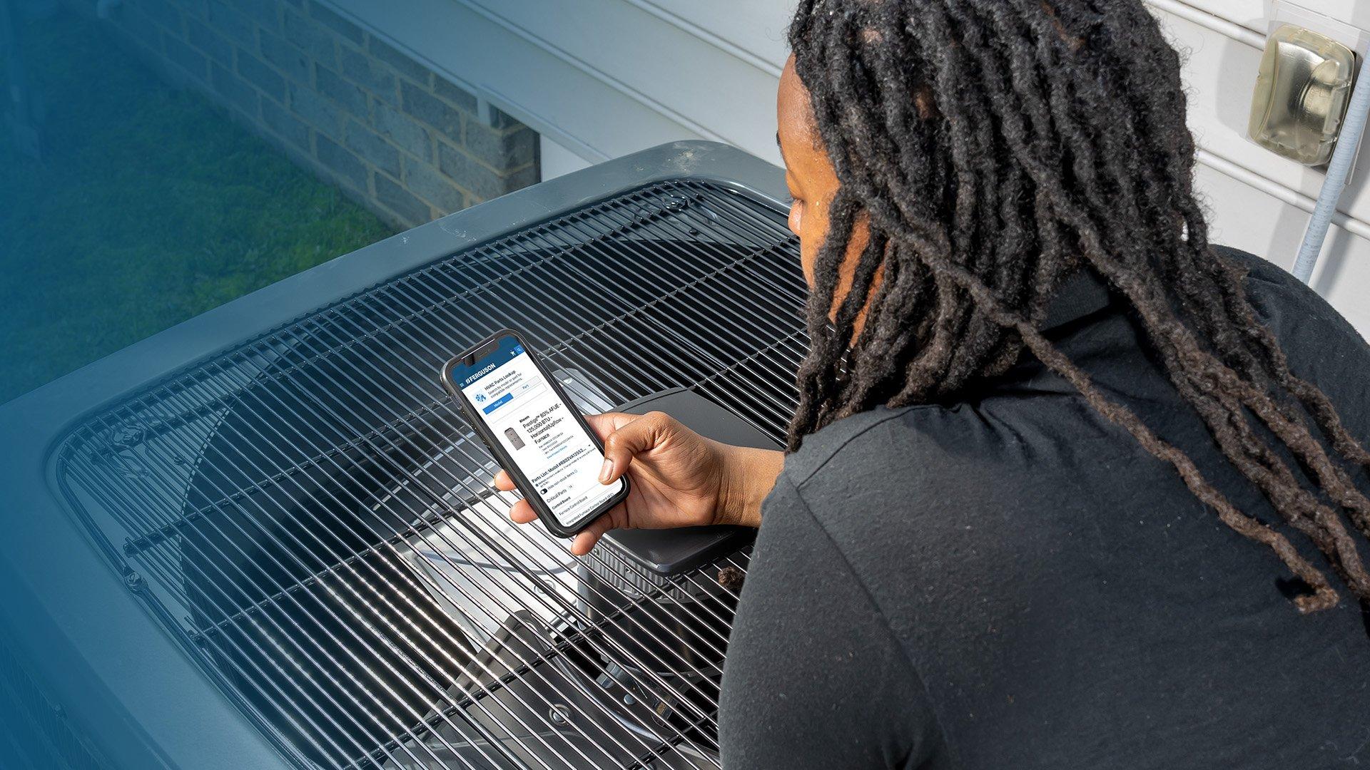 HVAC technician leans over HVAC unit while utilizing Ferguson's digital tools on smart phone. 