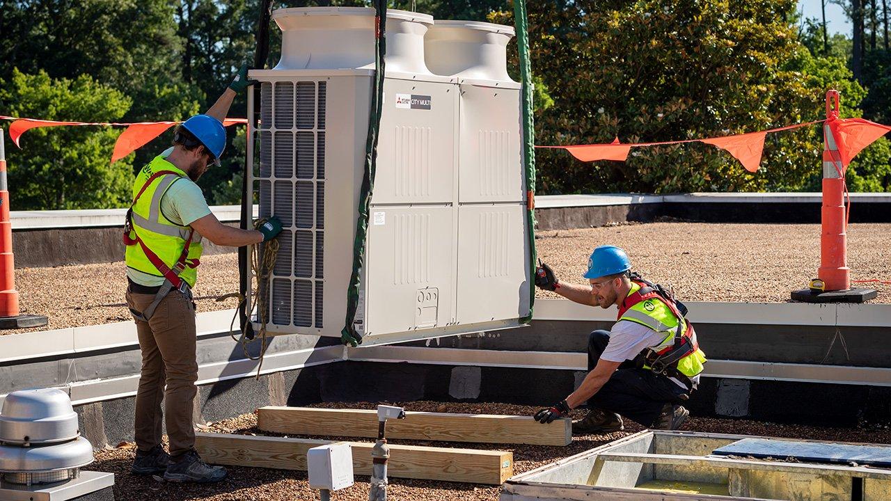 Two contractors guide a VRF outdoor unit into place on a rooftop.