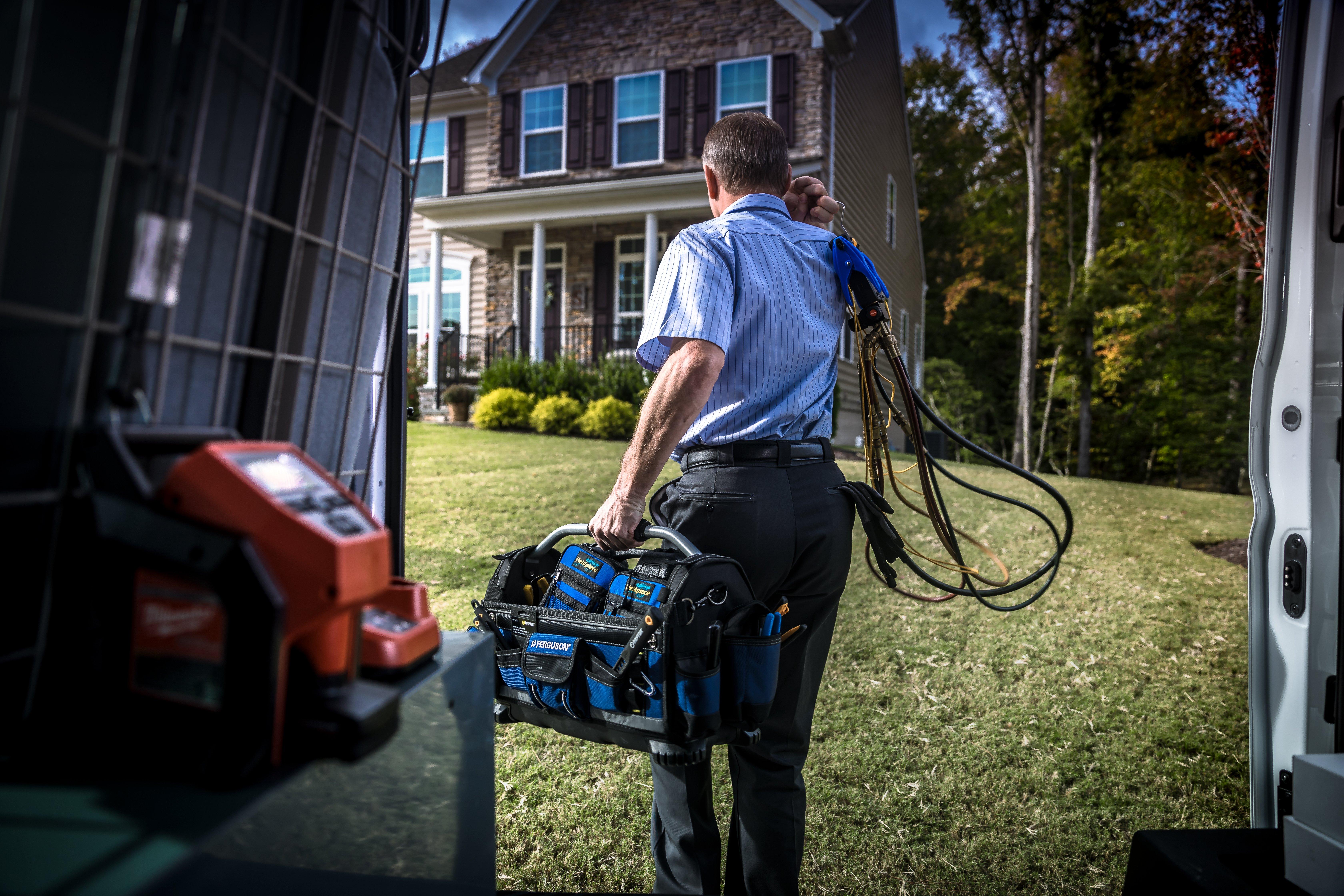 An HVAC technician carries a toolbag and cables from his work van toward a suburban two-story home.