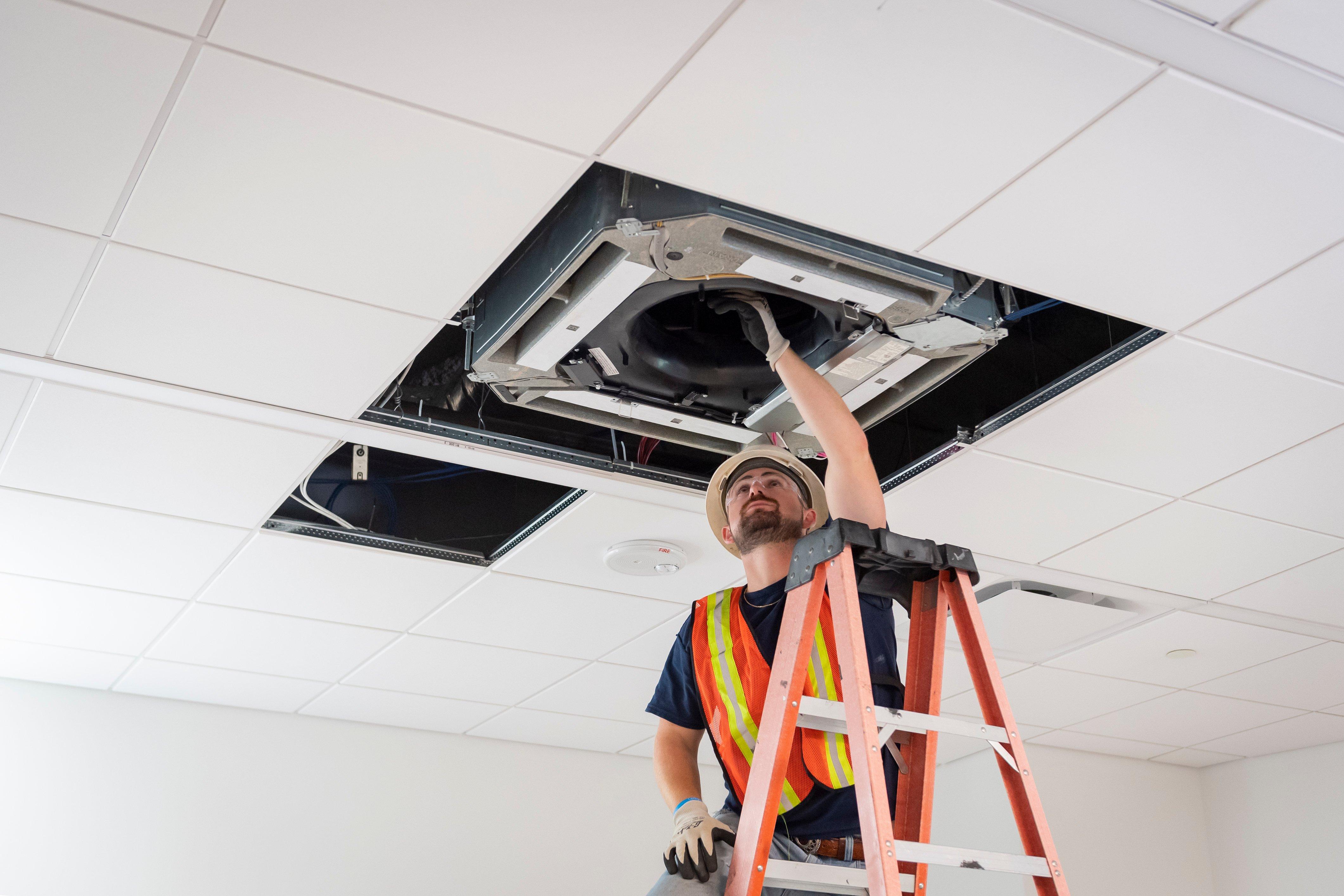 An HVAC technician on an orange ladder works on a ceiling cassette in a commercial building.